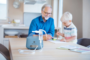 Opa spielt im Hintergrund mit Enkel am Tisch, im Vordergrund steht ein Inhalationsgerät mit Vernebler