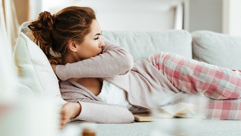 Junge Frau liegt auf der Couch und hustet in ihre Armbeuge