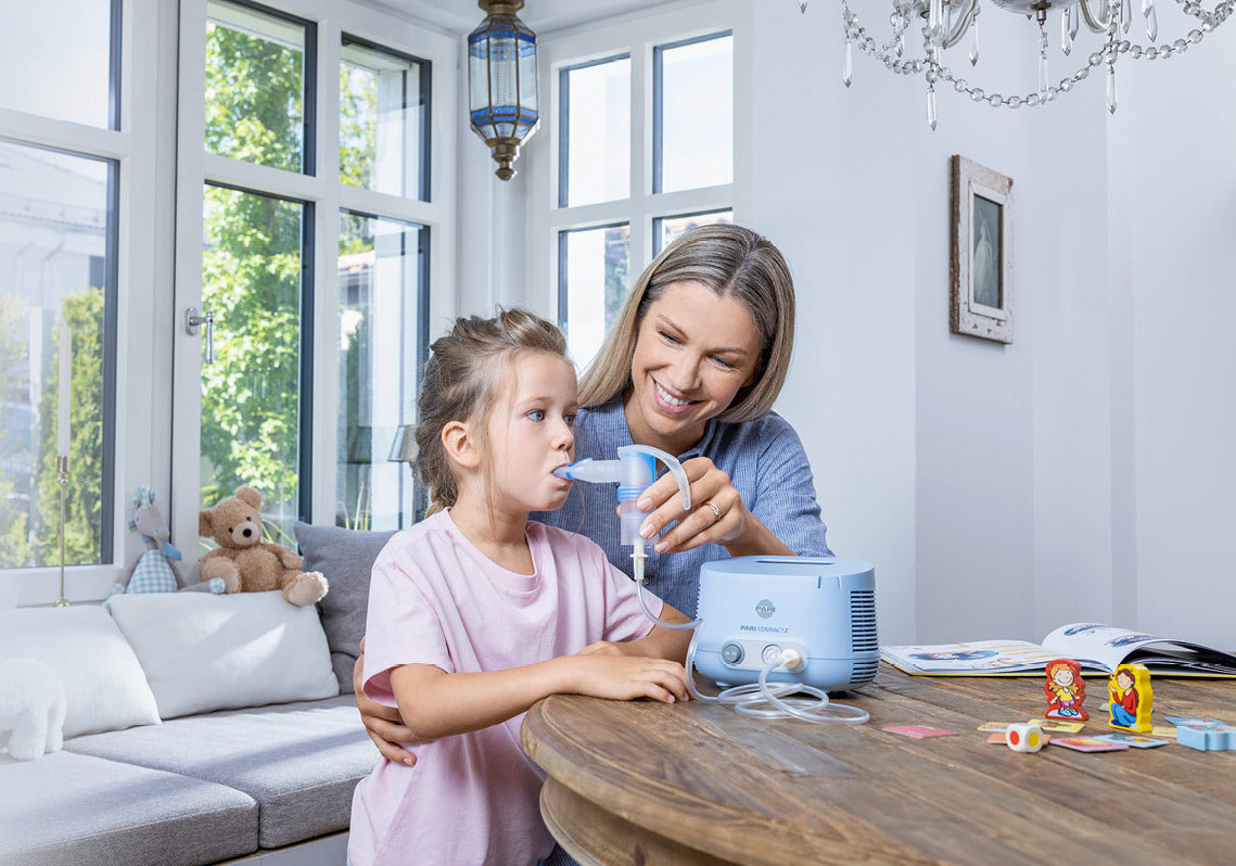 Eine Mutter hilft ihrer jungen Tochter beim Inhalieren mit dem PARI COMPACT2 Inhalationsgerät. Das Mädchen hält das Mundstück im Mund. Beide sitzen an einem runden Holztisch in einem hellen, freundlich eingerichteten Raum mit großen Fenstern.