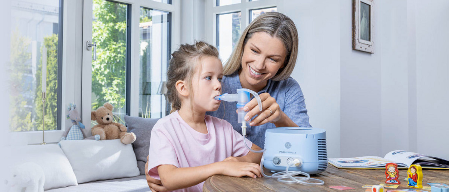 Eine Mutter hilft ihrer jungen Tochter beim Inhalieren mit dem PARI COMPACT2 Inhalationsgerät. Das Mädchen hält das Mundstück im Mund. Beide sitzen an einem runden Holztisch in einem hellen, freundlich eingerichteten Raum mit großen Fenstern.