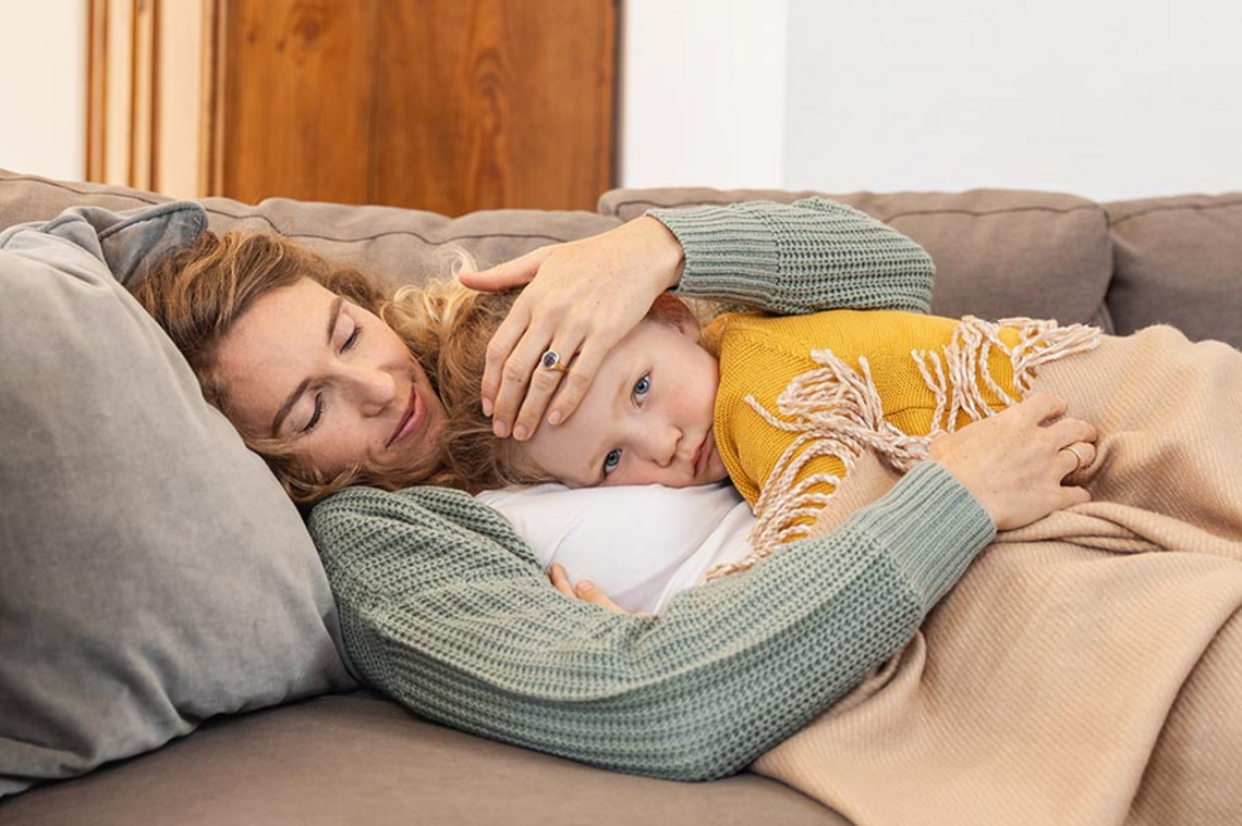 Symptome – So äußert sich das RS Virus Mutter liegt auf einem Sofa und hält ihr Kind im Arm, das auf ihrer Brust ruht, während sie dessen Stirn mit der Hand berührt.