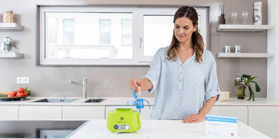 Eine Frau mit langen braunen Haaren steht in einer modernen, hellen Küche und füllt eine Inhalationslösung in den Vernebler des PARI BOY Junior, der vor ihr auf der Arbeitsplatte steht. Daneben steht eine Packung PARI Protect Inhalationslösung.