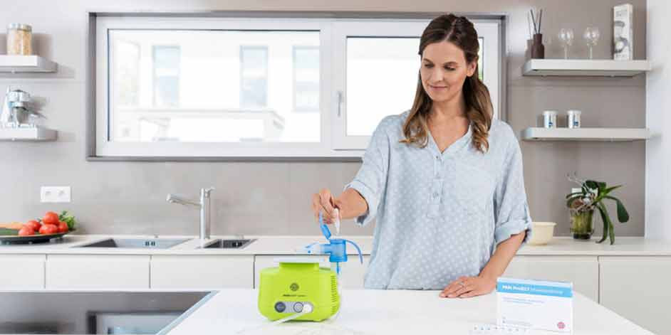 Eine Frau mit langen braunen Haaren steht in einer modernen, hellen Küche und füllt eine Inhalationslösung in den Vernebler des PARI BOY Junior, der vor ihr auf der Arbeitsplatte steht. Daneben steht eine Packung PARI Protect Inhalationslösung.