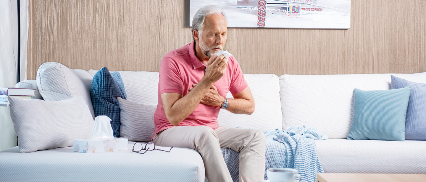 Älterer Mann sitzt auf einem Sofa, hält ein Taschentuch vor den Mund und eine Hand an die Brust, neben ihm liegen Taschentücher.