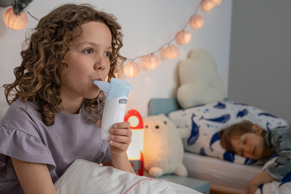 Husten beim Kind: PARI BOY® Go Kind sitzt im Bett und nutzt ein PARI BOY Go Inhalationsgerät mit Mundstück, weiteres Kind liegt im Hintergrund.