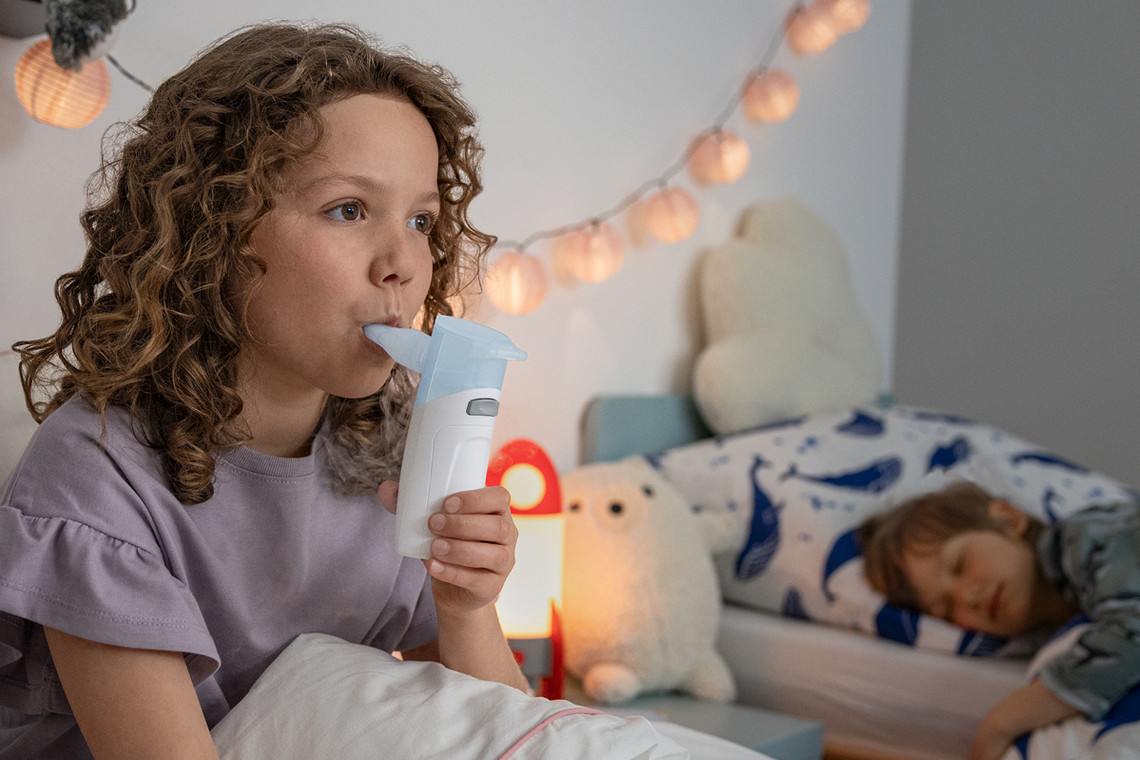 Richtig husten mit Kindern: Inhalieren Kind sitzt im Bett und nutzt ein PARI BOY Go Inhalationsgerät mit Mundstück, weiteres Kind liegt im Hintergrund.
