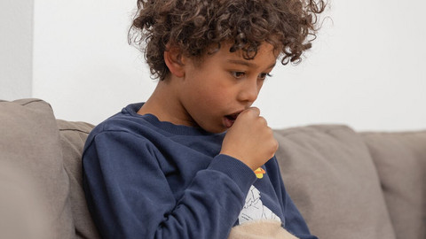 Junge sitzt auf dem Sofa und hält sich beim Husten eine Faust vor den Mund