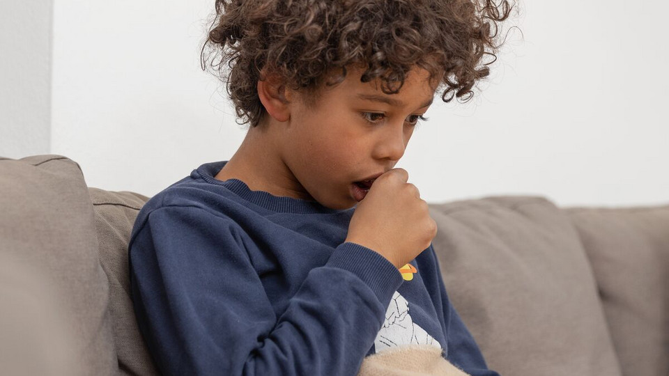Junge sitzt auf dem Sofa und hält sich beim Husten eine Faust vor den Mund