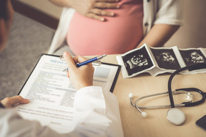 Hand einer Ärztin füllt einen Fragebogen aus, daneben Ultraschallbilder, Bauch einer Schwangeren im Hintergrund