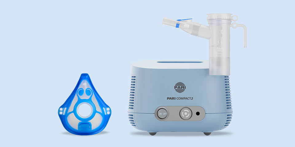 Produktabbildung: Vor einem blau-grauen Hintergrund steht ein PARI COMPACT2 Inhaliergerät mit aufgestecktem PARI LC PLUS Vernebler mit Mundstück. Links daneben liegt eine PARI Kindermaske soft.