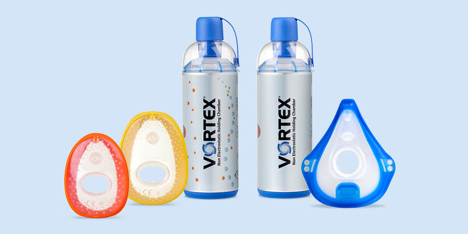 Produktabbildung – Im Hintergrund links steht eine VORTEX Inhalierhilfe für Kinder, rechts daneben die VORTEX für Erwachsene. Vor den Inhalierhilfen liegen von links nach rechts eine VORTEX Kindermaske, eine VORTEX Babymaske und die VORTEX Erwachsenenmaske.