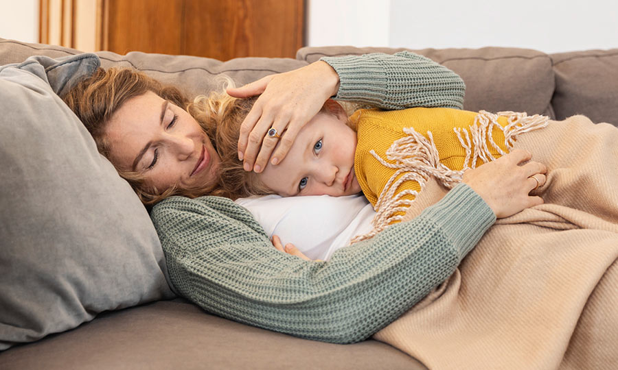 Mutter hält die Stirn ihres Kindes und liegt gemeinsam mit diesem auf dem Sofa 