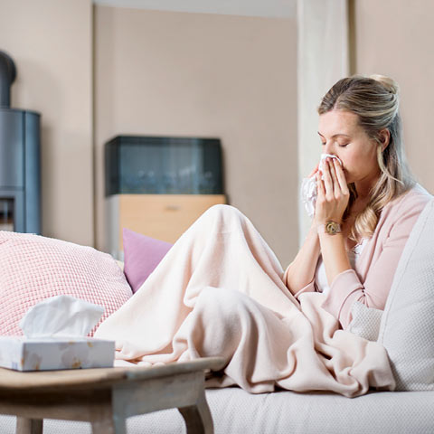 Eine Frau sitzt in eine Decke gehüllt auf einem Sofa und putzt sich mit einem Taschentuch die Nase. Sie sieht erschöpft aus. Auf dem Couchtisch vor ihr steht eine Box mit Papiertaschentüchern.