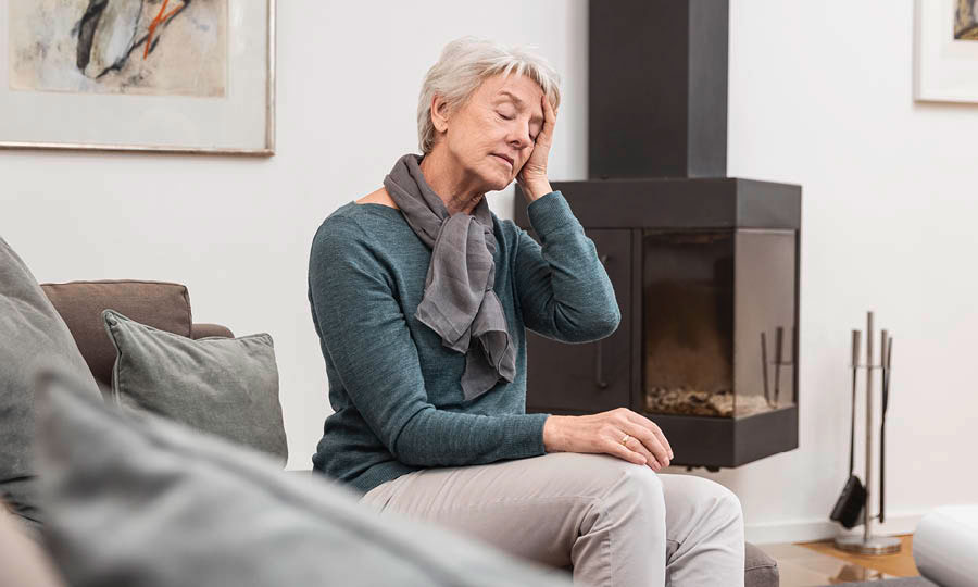 Person sitzt auf einem Sofa in einem modernen Wohnzimmer, trägt Schal und hält die Hand an die Schläfe, neben einem schwarzen Kamin.