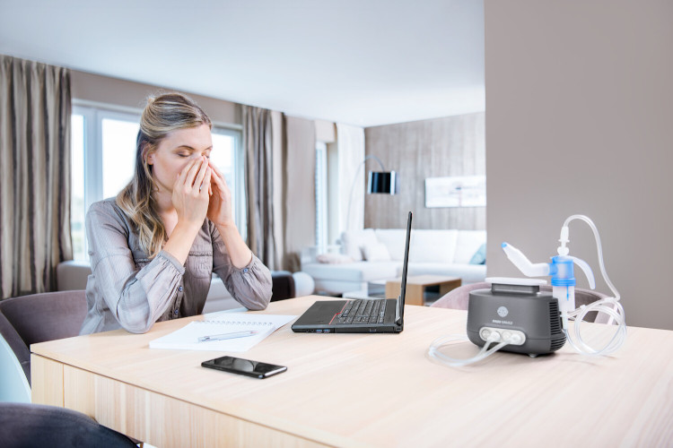 Woman sitting in front of laptop and holding both hands to her face, a nebuliser system can be seen in the foreground