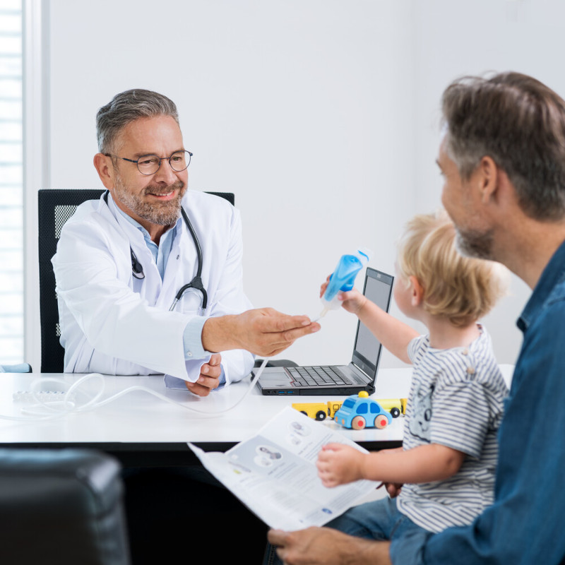 Vater sitzt mit Kind beim Arzt, der ihnen ein Inhalationsgerät zeigt