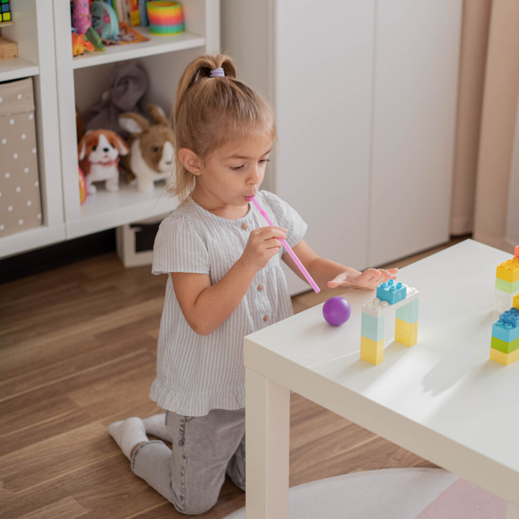 Kleines Mädchen kniet vor Spieltisch und bläst Ball mit Strohhalm durch kleine Duplo-Tore