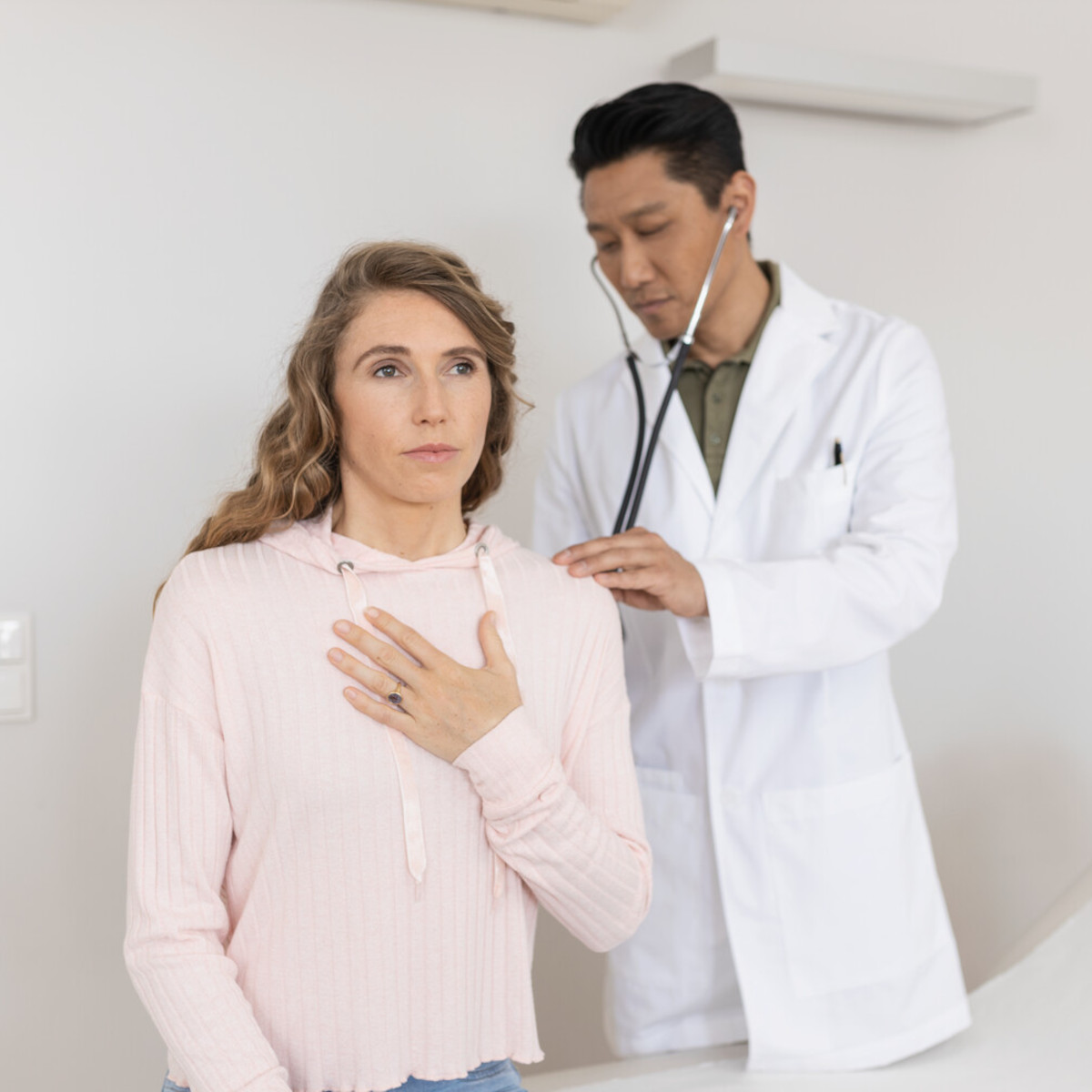 Junge Frau sitzt auf einer Liege mit Hand auf der Lunge, während Arzt hinter ihr steht und sie mit Stethoskop abhört