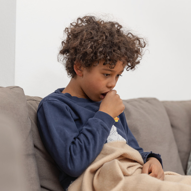 Junge sitzt hustend auf dem Sofa, er hält sich eine Hand vor den Mund