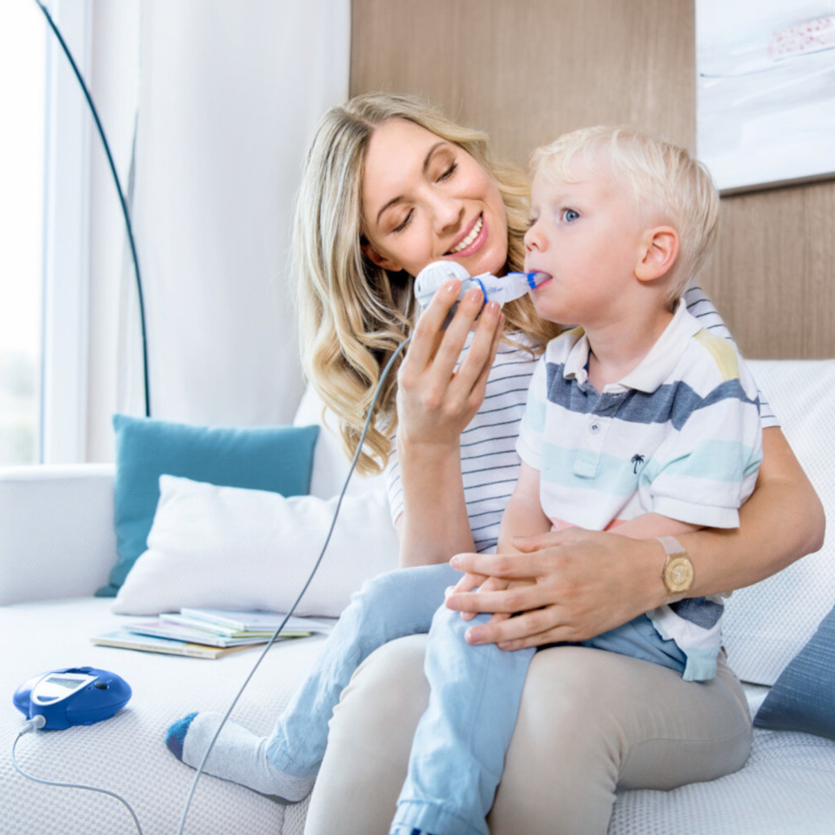 Mutter sitzt mit ihrem Kind auf dem Schoß auf der Couch und hilft ihm beim Inhalieren mit dem eFlow rapid Inhalationsgerät für Mukoviszidose