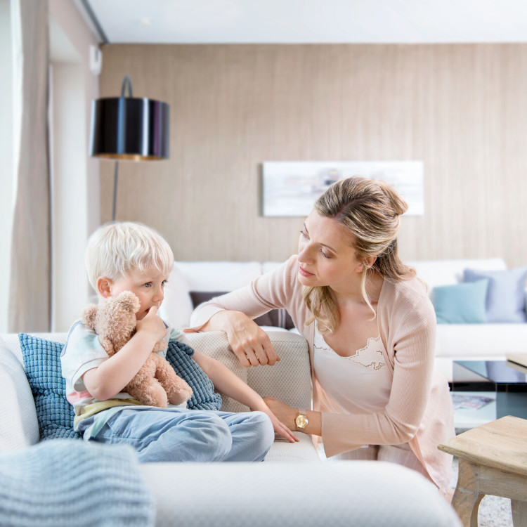 Kleiner Junge sitzt mit Kuscheltier auf der Couch, Mutter sitzt besorgt davor