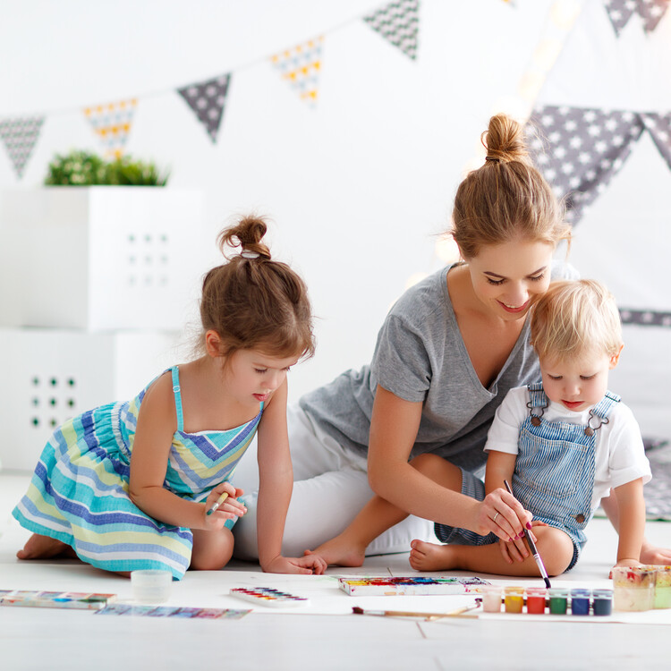 Mutter sitzt mit zwei kleinen Kindern im Kinderzimmer am Boden und malt mit Wasserfarben