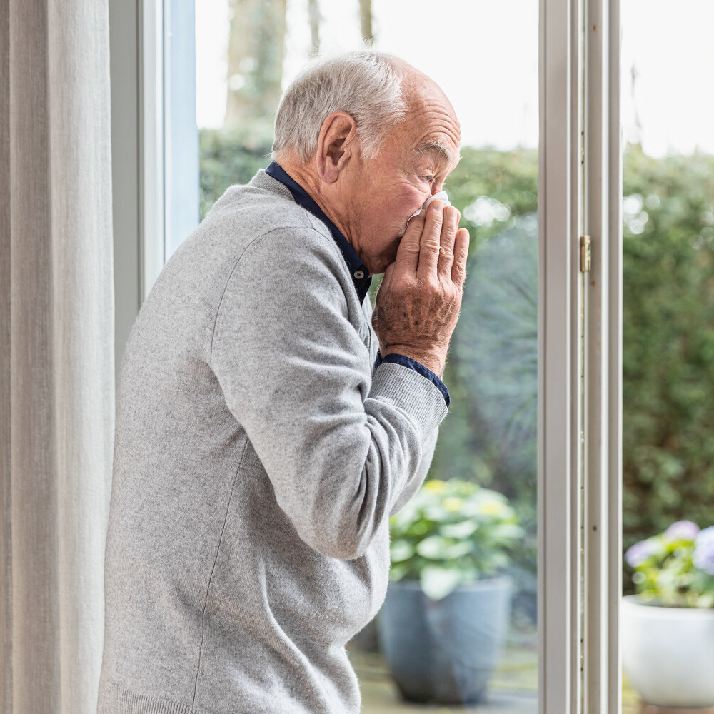 Älterer Mann steht vor der geöffneten Terrassentür und niest in ein Taschentuch