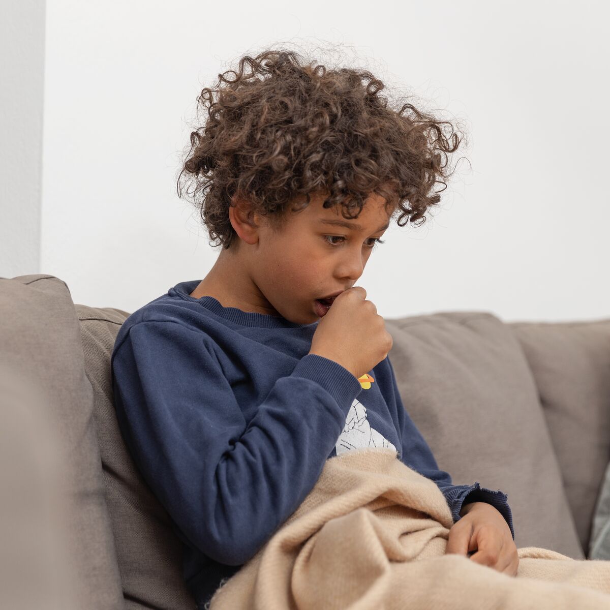Junge sitzt auf der Couch mit einer Decke, er hustet und hält sich eine Faust vor den Mund