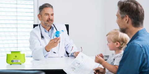 Vater mit Sohn auf seinem Schoß sitzend beim Arzt, welcher beiden die Funktionsweise eines Verneblers beschreibt