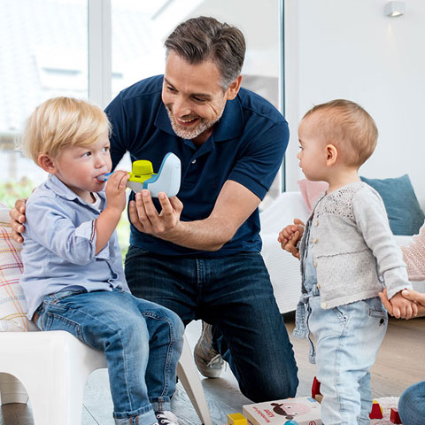 Ein Vater hilft seinem kleinen Jungen bei der Benutzung eines Inhalationsgeräts, während die Mutter die Hand eines zweiten jüngeren Kindes hält und lächelnd zusieht.