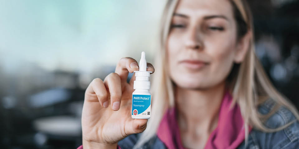 Person hält eine 20 ml Flasche PARI Protect Nasenspray mit weiß-blauem Etikett vor verschwommenem Hintergrund in der Hand.