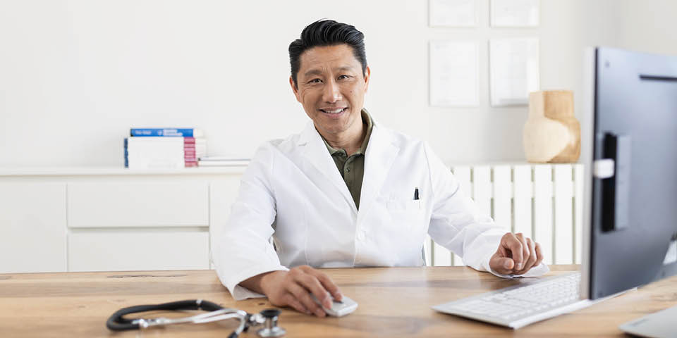 Arzt in weißem Arztkittel sitzt am Schreibtisch vor einem Computer, daneben liegt ein Stethoskop.