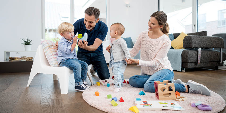 Zwei Erwachsene spielen mit zwei Kleinkindern auf einem Teppich, umgeben von buntem Spielzeug und Holzformen.