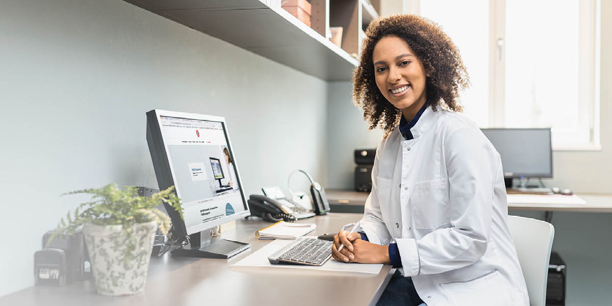 Person in weißem Kittel sitzt am Schreibtisch und arbeitet an einem Computer, daneben Telefon und Pflanze.