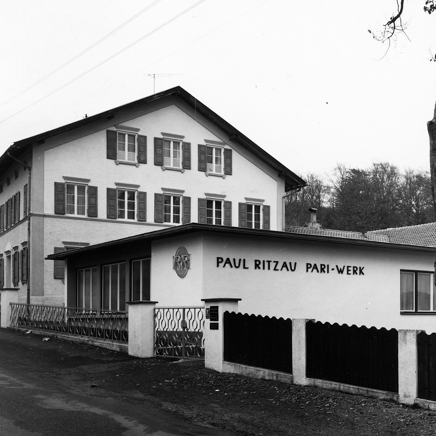 1906 Firmengründung – Mehrstöckiges Gebäude mit Giebeldach und Schild „PAUL RITZAU PARI-WERK“, umgeben von Zaun und Bäumen im Hintergrund.