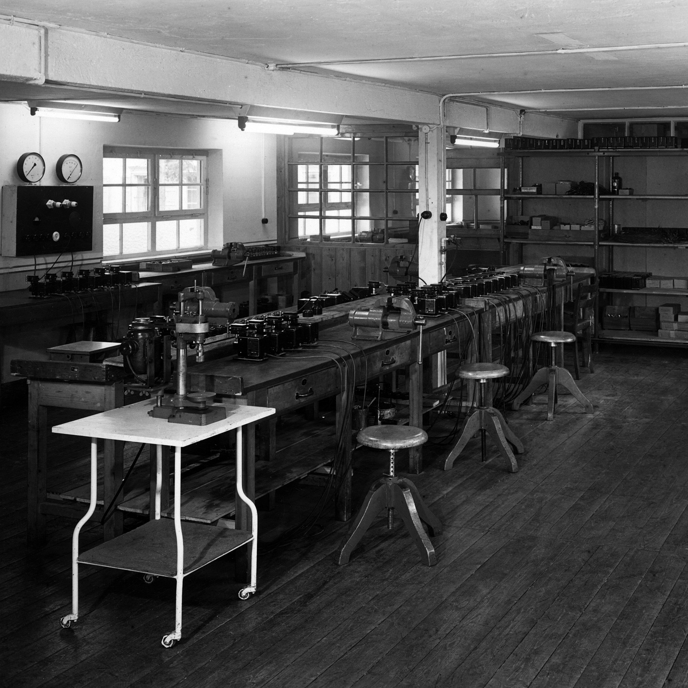 A black-and-white photo of an old laboratory with long workbenches, technical equipment, cables, and stools in a workspace.