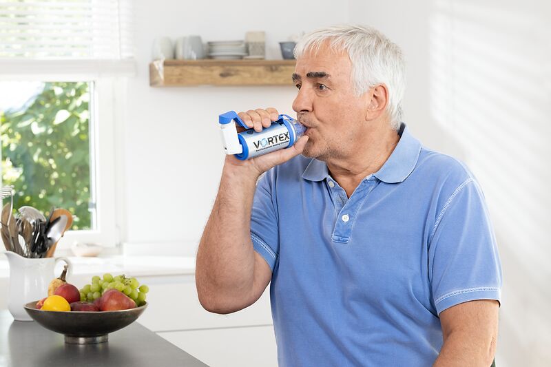 Elderly man uses inhaler with VORTEX spacer in bright kitchen, fruit bowl and kitchen utensils in the background.
