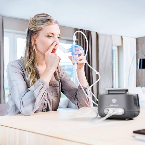 A woman uses the PARI SINUS2 inhaler for sinus therapy. She holds the mouthpiece with one hand and closes one nostril with the other. The device is placed on a table.