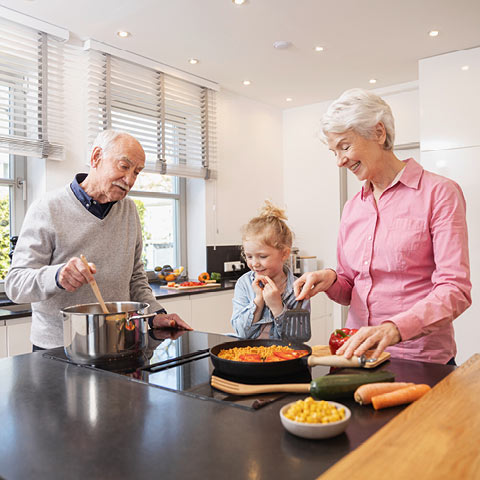 An elderly couple cooks together with their grandchild in a kitchen. The grandmother chops vegetables while the grandfather stirs the stove.