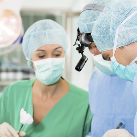 A team of surgeons in scrubs performs an operation. One doctor wears magnifying glasses with a light, while a resident holds sterile instruments.