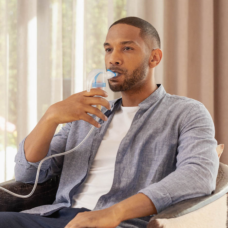 The person is sitting at home and using an inhalation device with a mouthpiece for respiratory therapy.