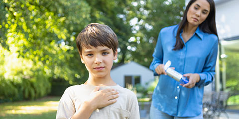 A boy stands in the garden with one hand on his chest, while a woman in a blue blouse stands worriedly behind him with a VORTEX inhaler in her hand.