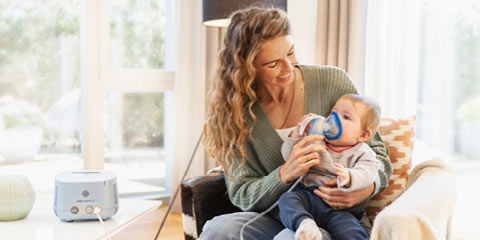 A smiling woman with long, wavy hair holds her baby on her lap and helps him use an inhalation mask.