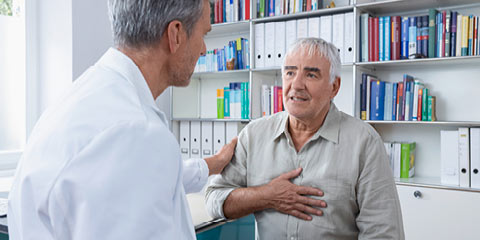 An elderly man sits in a doctor's office, holds his hand on his chest and talks to a doctor in a white coat who places a reassuring hand on his shoulder.