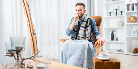 A young man wearing a scarf and blanket sits on an armchair in a cozy living room and coughs.