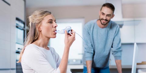 A woman sits in a modern kitchen using an inhaler while a man watches her, smiling and leaning on the countertop.