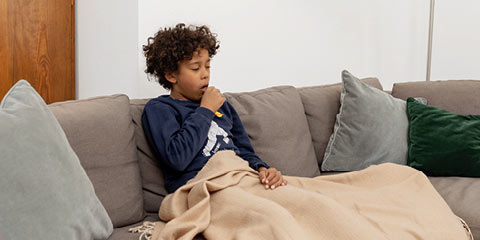A boy with curly hair sits sick on a gray sofa, wrapped in a beige blanket. He coughs into his hand.