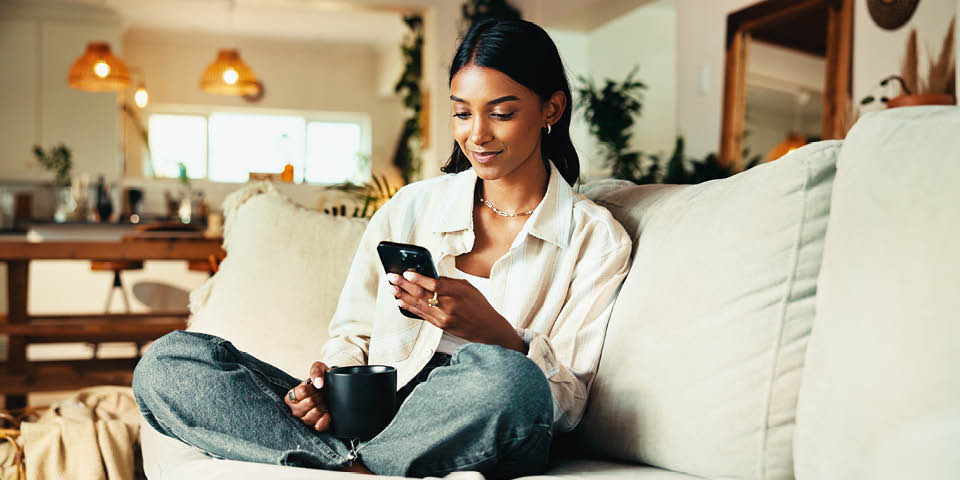 A person sits relaxed on the sofa, holding a smartphone and a cup in the living room.