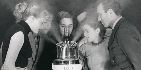 Old black and white photograph: A group of five people stand around an inhalation device from which thick vapour is flowing. They all look at the device with fascination and breathe in the aerosol.