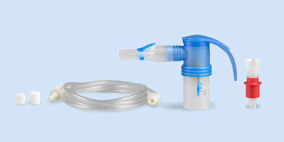 Imagem do produto: Num fundo azul-acinzentado, encontra-se no centro um nebulizador PARI LC SPRINT Junior com bocal. À direita, um bocal vermelho. À esquerda, encontra-se a mangueira de ligação, ao lado de dois filtros diferentes para o compressor.
